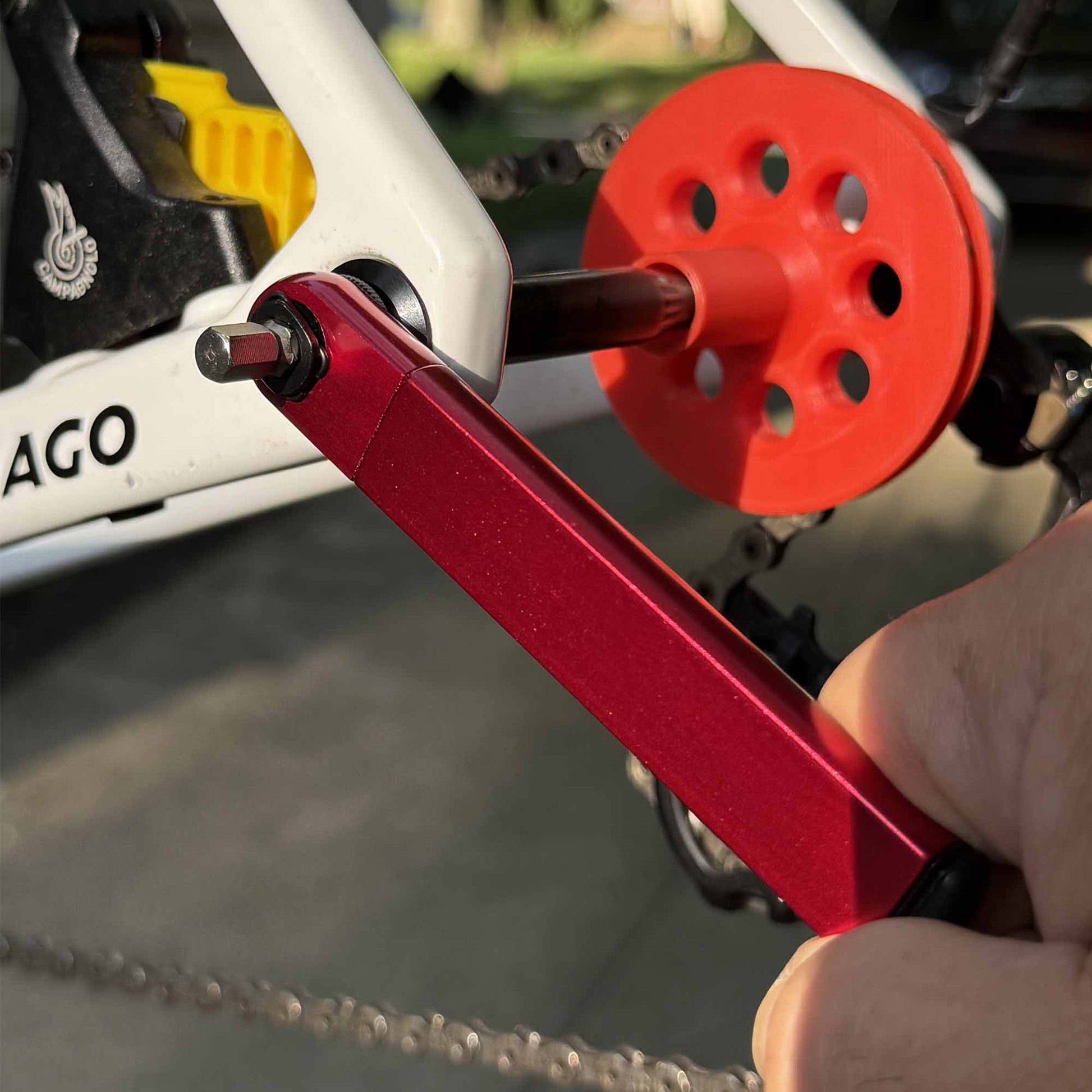A close-up of a hand using the Prestacycle PocketRatchet Lite, a pocket multi-tool with bits stored in the handle, to adjust the crankset bolt on a white bicycle frame, with a red plastic wheel near the rear derailleur.